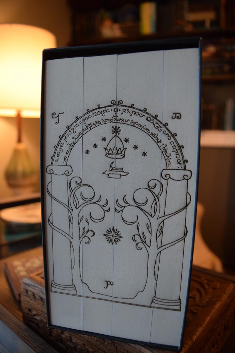 May include: A black and white illustration of a door with an archway, crown, and trees. The design is reminiscent of a fantasy theme. The image is on a light-colored surface, possibly a book cover or box.