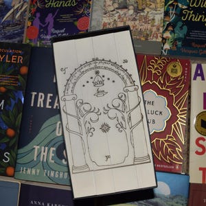 May include: A white and black decorative bookmark featuring an arched doorway design with trees and a crown. The bookmark is rectangular and rests on a collection of books with colorful covers, including "Light From Other Stars."