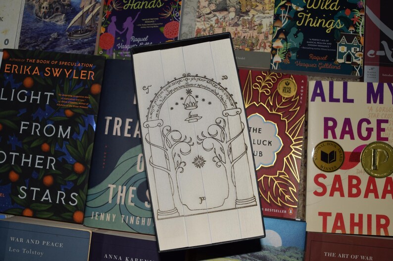 May include: A white and black decorative bookmark featuring an arched doorway design with trees and a crown. The bookmark is rectangular and rests on a collection of books with colorful covers, including "Light From Other Stars."