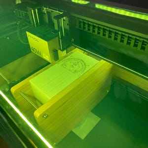 May include: An XTOOL 40W laser engraver with a stack of light green paper inside. The machine is enclosed in a dark frame with a green glow. The paper has a design etched into the top sheet.