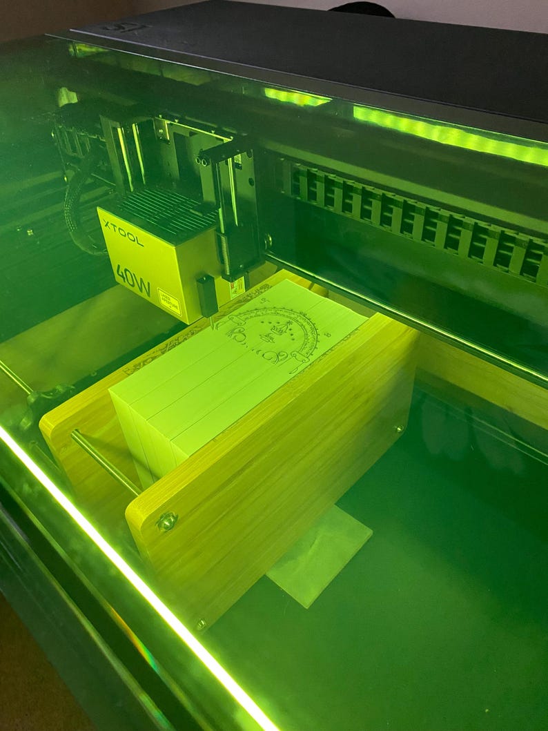 May include: An XTOOL 40W laser engraver with a stack of light green paper inside. The machine is enclosed in a dark frame with a green glow. The paper has a design etched into the top sheet.
