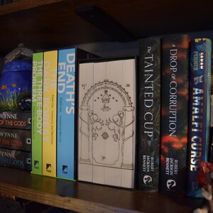 May include: A bookshelf displays a collection of books with colorful spines. Titles include "Death's End," "The Three-Body Problem," and "The Tainted Cup." A decorative teapot and a small figurine are also visible on the shelf.
