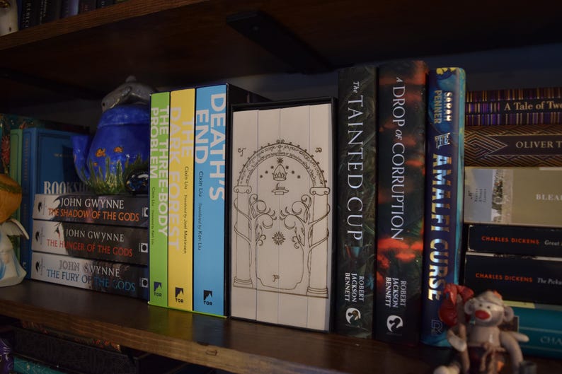 May include: A bookshelf displays a collection of books with colorful spines. Titles include "Death's End," "The Three-Body Problem," and "The Tainted Cup." A decorative teapot and a small figurine are also visible on the shelf.