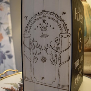 May include: A boxed set of "The Hobbit" and "The Lord of the Rings" books. The spine features a detailed illustration of a doorway with trees and an archway. The book's title is in gold lettering on a black background.