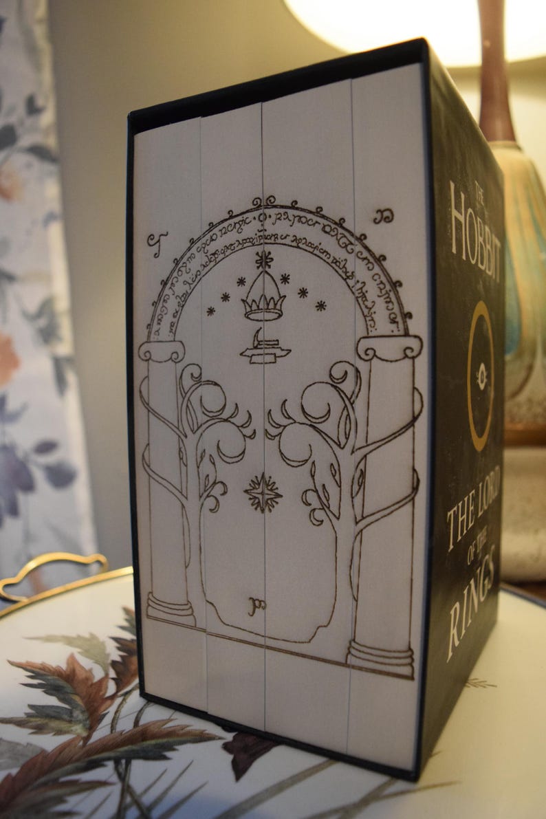May include: A boxed set of "The Hobbit" and "The Lord of the Rings" books. The spine features a detailed illustration of a doorway with trees and an archway. The book's title is in gold lettering on a black background.