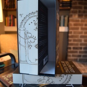 May include: A boxed set of "The Fellowship of the Ring" book. The book is in a decorative box with gold-colored line art and text. The book's spine is visible, with the title printed in gold. The box is open, revealing the book inside.