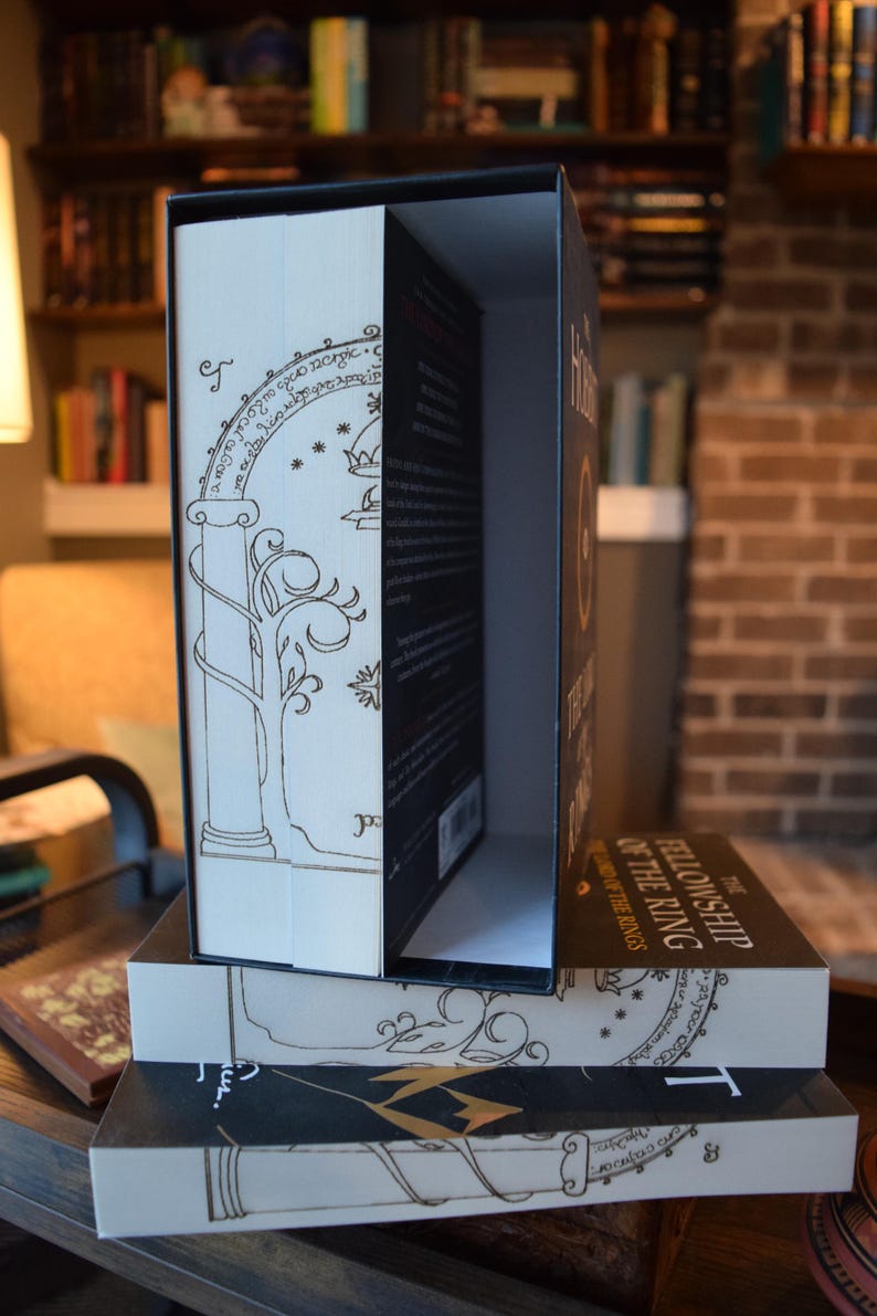 May include: A boxed set of "The Fellowship of the Ring" book. The book is in a decorative box with gold-colored line art and text. The book's spine is visible, with the title printed in gold. The box is open, revealing the book inside.