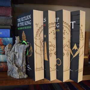 May include: A collection of books, including "The Return of the King" by J.R.R. Tolkien, displayed on a wooden shelf. The books have black spines with gold lettering and illustrations. A statue of a figure stands in front of the books.