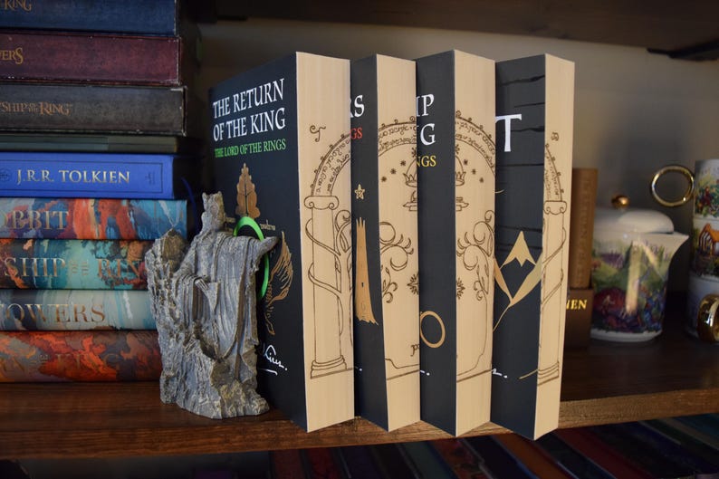 May include: A collection of books, including "The Return of the King" by J.R.R. Tolkien, displayed on a wooden shelf. The books have black spines with gold lettering and illustrations. A statue of a figure stands in front of the books.