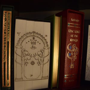 May include: A collection of books, including "The Hobbit" and "The Lord of the Rings", displayed on a shelf. The books have decorative covers in green and red, with gold lettering and illustrations. The image evokes a fantasy theme.