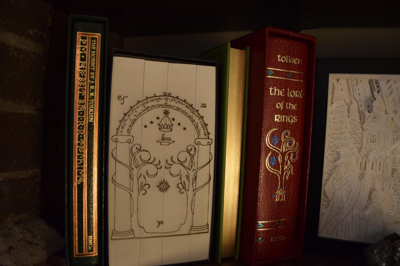 May include: A collection of books, including "The Hobbit" and "The Lord of the Rings", displayed on a shelf. The books have decorative covers in green and red, with gold lettering and illustrations. The image evokes a fantasy theme.