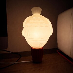 Könnte beinhalten: Dekorative Lampe in Fischform, mit einem leuchtenden weißen Körper und einem roten Sockel. Die Lampe besteht aus einem durchscheinenden Material, und das Fischdesign umfasst Flossen, Augen und Schuppen. Die Lampe steht auf einer Holzoberfläche.