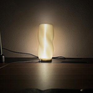 Könnte beinhalten: Eine moderne, beleuchtete Tischlampe mit einem einzigartigen, gerippten Design. Die Lampe strahlt ein warmes, weiches Licht aus und schafft eine gemütliche Atmosphäre. Links befindet sich eine schwarze Schreibtischlampe. Die Lampe steht auf einer dunklen Holzoberfläche.