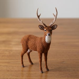 May include: A needle-felted deer figurine with a textured brown coat, a white patch on the chest, and detailed antlers. The deer stands in a forward-facing position with dark eyes and pointed ears, showcasing a soft, fuzzy appearance throughout.