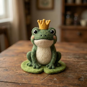 May include: A needle-felted green frog figurine with a cream-coloured belly and pink cheeks, wearing a small yellow crown on its head. The frog is sitting on a flat, circular green lily pad base, featuring large black eyes and a simple stitched mouth.
