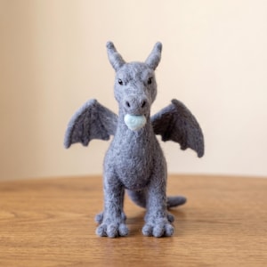 May include: A needle-felted grey dragon figurine with two wings, pointed ears, and small black eyes. The dragon has a light blue circular object held in its mouth. The creature stands on four legs with visible claws and has a textured, fuzzy surface.