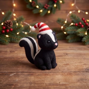 Op de afbeelding: Een naaldgevilte stinkdierfiguur met zwart-witte vacht, een witte streep van de kop tot de rug en een pluizige staart. Het stinkdier draagt een klein, zacht rood-wit gestreept kerstmutsje met een wit pomponnetje aan de bovenkant.