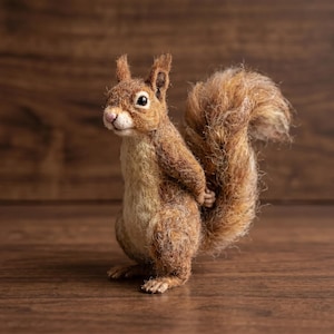 Peut inclure: Une figurine d'écureuil en laine feutrée à l'aiguille, composée de fibres brunes et crème texturées. L'écureuil se tient debout, les pattes avant repliées derrière le dos, avec une queue touffue, des oreilles pointues et un petit nez rose.