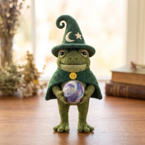 May include: A needle-felted green frog figurine wearing a dark green wizard hat with a white crescent moon and star, and a matching cape fastened with a yellow button. The frog holds a small, round, swirled purple, blue, and white wool orb.