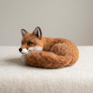 May include: A needle-felted fox figurine in a curled sleeping position. The fox features textured orange-brown fur, a white muzzle, black-tipped ears, and a black nose. The body is coiled with the tail wrapped around the front of the torso.