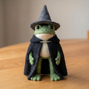 May include: A handcrafted felt frog figurine wearing a gray wizard hat and a black cape. The frog has green skin, a white belly, and is standing on a wooden surface. The cape is tied with a string.