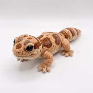Könnte beinhalten: Eine nadelgefilzte Leopardgecko-Figur aus beigefarbener und brauner Wolle. Die Eidechse hat eine strukturierte, flauschige Oberfläche, dunkelbraune Flecken auf dem Rücken, einen dicken, segmentierten Schwanz und zwei große, glänzende schwarze Perlenaugen.