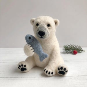May include: A needle-felted polar bear figurine in off-white wool, featuring a grey muzzle, black nose, and dark eyes. The bear is sitting upright and holding a small, light blue felted fish in its front paws. It has black felted paw pads.