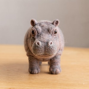 Puede incluir: Una figura de hipopótamo hecha con fieltro de aguja, elaborada con fibras de lana texturizadas en tonos gris y marrón jaspeado. La pequeña escultura animal presenta orejas redondeadas, un hocico ancho con fosas nasales y dos ojos oscuros.