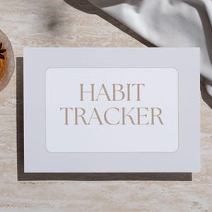 Puede incluir: Una tarjeta rectangular blanca con las palabras "HABIT TRACKER" en una fuente marrón claro. Un vaso de líquido ámbar con anís estrellado está en la esquina superior izquierda. Un paño blanco está en la esquina superior derecha.