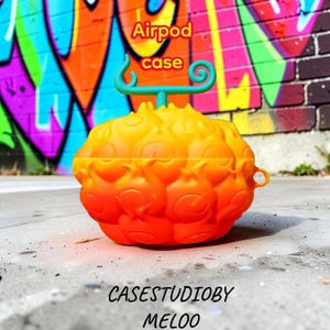 May include: Orange and yellow Airpod case shaped like a fruit, with a green stem and swirl design. The case has a textured surface and the words "Airpod case" are written in red and yellow. The text "CASESTUDIOBY MELOO" is written in white.