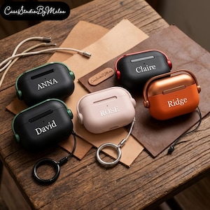 Könnte beinhalten: Personalisierte AirPods-Hüllen in verschiedenen Farben, darunter Schwarz, Rosa und Orange. Jede Hülle ist mit einem Namen in weißer Schrift graviert. Die Hüllen werden auf einer Holzoberfläche mit Lederakzenten präsentiert.