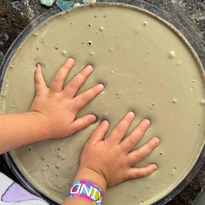 May include: A circular, clear plastic tray filled with a smooth, light gray, viscous substance featuring small air bubbles throughout. A colorful wristband with the text KIND is visible on one wrist, and scattered blue and teal sea glass pieces.