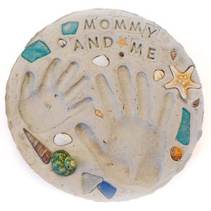 May include: A round, light gray concrete stepping stone with handprints and the words "MOMMY AND ME." The stone is decorated with seashells, a starfish, and pieces of blue and green glass. A unique keepsake.