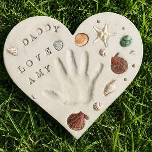 May include: A heart-shaped light gray stone plaque featuring an imprinted child's handprint in the center. The text DADDY LOVE AMY is embossed on the left side. The surface is decorated with various seashells, a starfish, and a small blue heart.