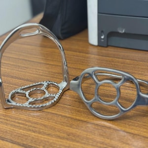 Op de afbeelding: Twee zilverkleurige paardentrappen. De stijgbeugels hebben een gebogen bovenkant en een voetsteun met een open ontwerp. De voetsteunen hebben cirkelvormige en ovale uitsparingen. De stijgbeugels zijn gemaakt van glanzend metaal.
