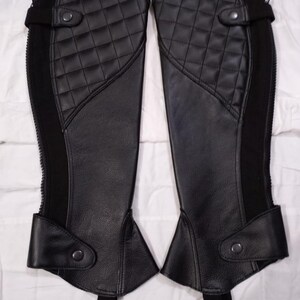 May include: A pair of black equestrian half chaps featuring a quilted upper panel, a smooth lower section, and a vertical side zipper. Each chap includes a snap button closure at the top and bottom, along with an elastic stirrup strap at the base.
