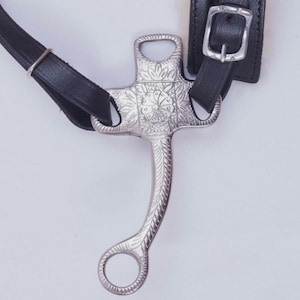 May include: A detailed image of a silver horse bit featuring floral patterns. The bit is attached to black leather straps, secured with a silver buckle. The bit has a curved shape and a ring at the base.