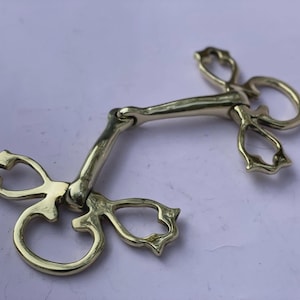 Op de afbeelding: Een goudkleurige paardenbit met decoratieve hartvormige en gebogen accenten. De bit heeft een centrale stang en ringen aan weerszijden voor de teugels. Het metaal heeft een gepolijste afwerking.