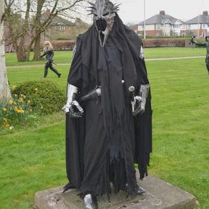 May include: A figure in a black costume with a tall, spiked helmet and a long, tattered cloak stands on a stone base in a park. The costume includes silver gauntlets and boots. Other costumed figures are in the background.