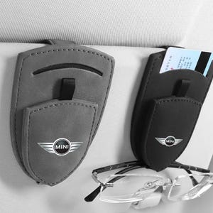 Puede incluir: Dos clips para visera de coche en forma de escudo, en gris y negro, con el logotipo de MINI. El clip gris tiene una ranura horizontal, mientras que el negro sujeta una tarjeta blanca y unas gafas sin montura colgadas de una cinta negra.
