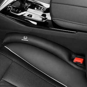 以下が含まれることがあります： 表面に白いホンダのロゴがプリントされた黒いレザー製のカーシート隙間埋めクッション。このアクセサリーは、車両のシートとセンターコンソールの間に配置され、隙間に物が落ちるのを防ぐために使用されます。