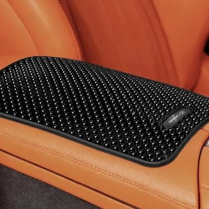 May include: A black rectangular car center console armrest cover featuring a textured surface with numerous small raised bumps. The cover is positioned on an orange leather armrest and includes a small black rectangular logo emblem on the right side.