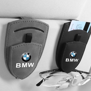 May include: Two car visor organizers, one gray and one black, featuring the BMW logo and text. Each organizer has a shield-shaped front flap, a small strap, and a slot for cards. The black organizer holds a pair of rimless eyeglasses on its clip.