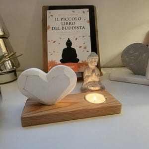 Può includere: Un portacandele in legno con una candela accesa, una scultura a forma di cuore bianco e una piccola statua di Buddha. Un libro intitolato "Il Piccolo Libro Del Buddista" è sullo sfondo. Gli oggetti sono disposti su una superficie bianca.