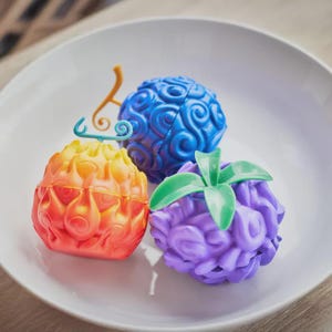 May include: Three colorful, stylized fruit-shaped objects. One is blue, one is orange and yellow, and the third is purple. Each has a unique textured surface and a stem or leaf detail. They are arranged on a white plate.