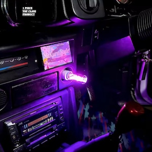 May include: Interior car detail with a purple glow. A light is plugged into the cigarette lighter. The car stereo is visible, along with the text "Sakura Style" on a card. The text "1 Piece Top Class Product" is also visible.
