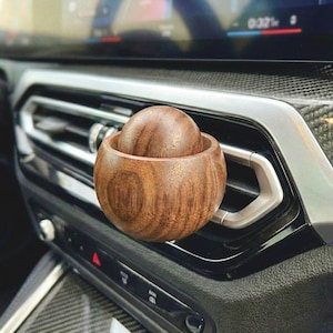 Puede incluir: Un difusor de ventilación de coche de madera con un diseño esférico. El difusor está hecho de madera marrón oscuro y tiene una esfera de madera a juego en el interior. El producto está diseñado para engancharse a la ventilación del coche.