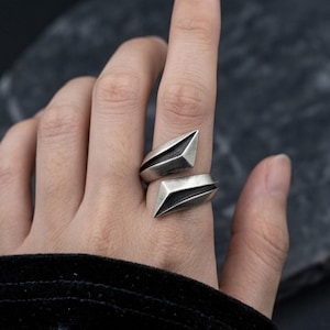 May include: A silver-toned open ring featuring two angular, geometric, pointed ends that overlap slightly. The metal has a matte, oxidized finish with deep shadows in the recessed areas, creating a sharp, architectural look on the finger.