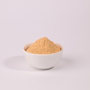 May include: A white ceramic bowl filled with a light brown powder, possibly a spice or flour, sits against a plain white background. The powder is finely ground and has a slightly textured appearance. The bowl is round and simple in design.