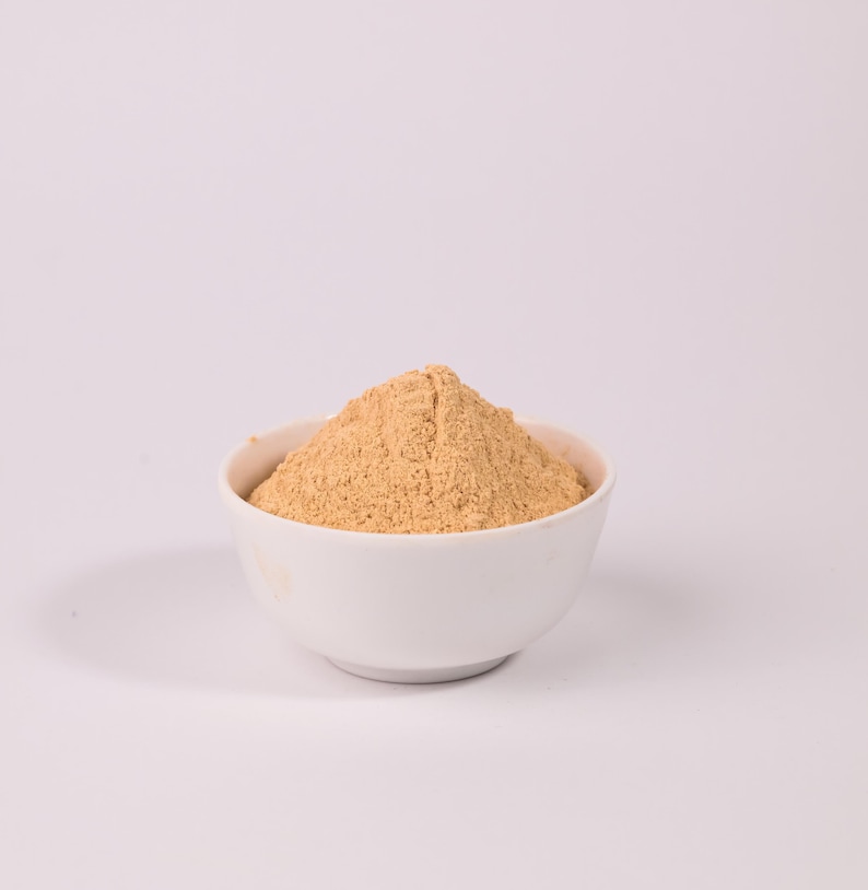 May include: A white ceramic bowl filled with a light brown powder, possibly a spice or flour, sits against a plain white background. The powder is finely ground and has a slightly textured appearance. The bowl is round and simple in design.